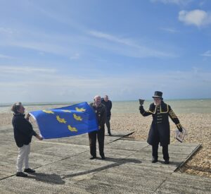 The Mayor, Town Crier of Seaford and guest holding out the Sussex Day flag at Seaford Beach